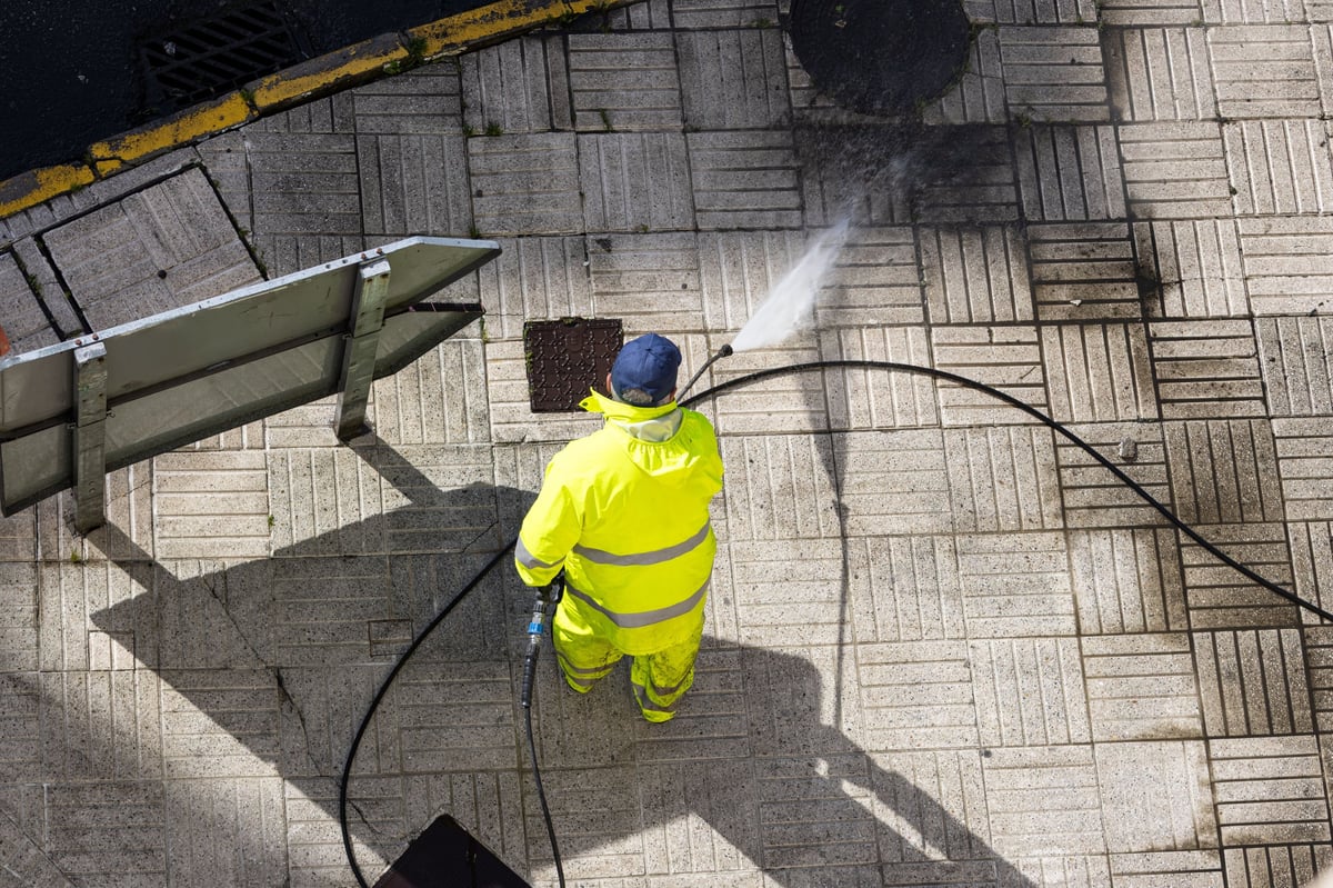 A worker uses a high pressure water jet machine to clean a street sidewalk and exterior surface.
